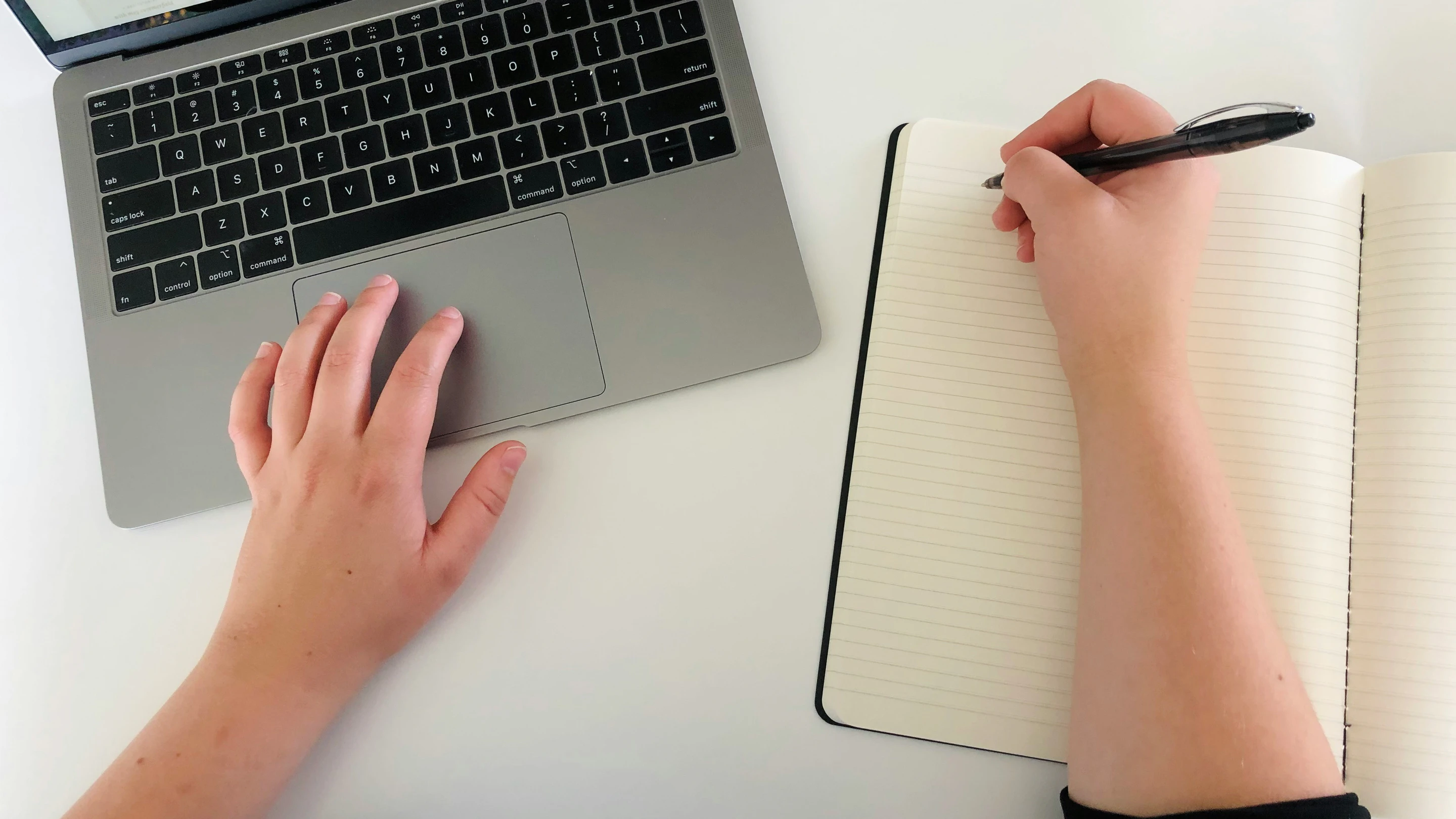 Overhead view of a person working at a white desk, with their left hand resting on a laptop trackpad and right hand holding a pen over an open lined notebook.